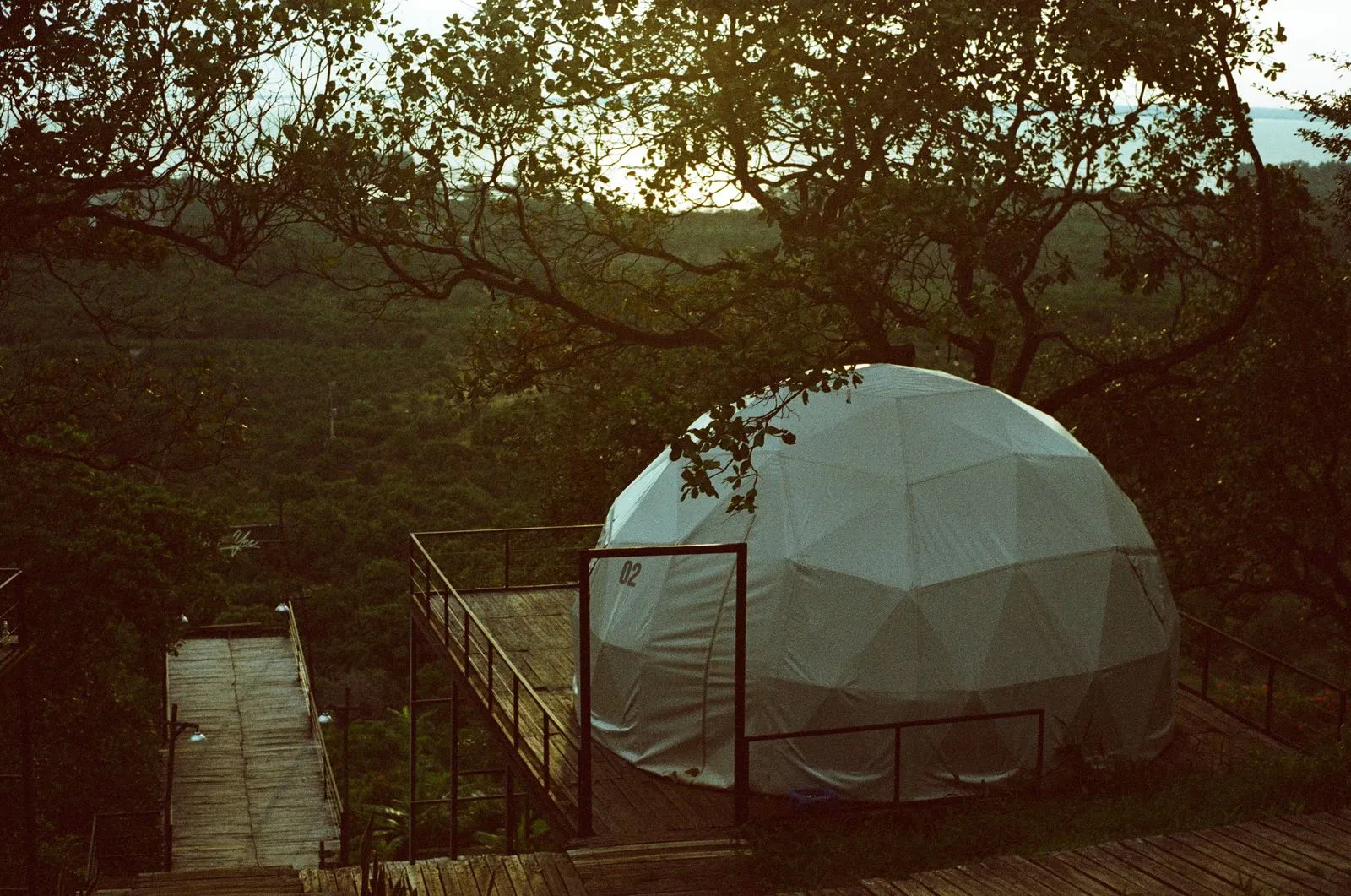 Dom geodezic de glamping pe o terasă de lemn, la apus, cu vedere peste dealuri împădurite