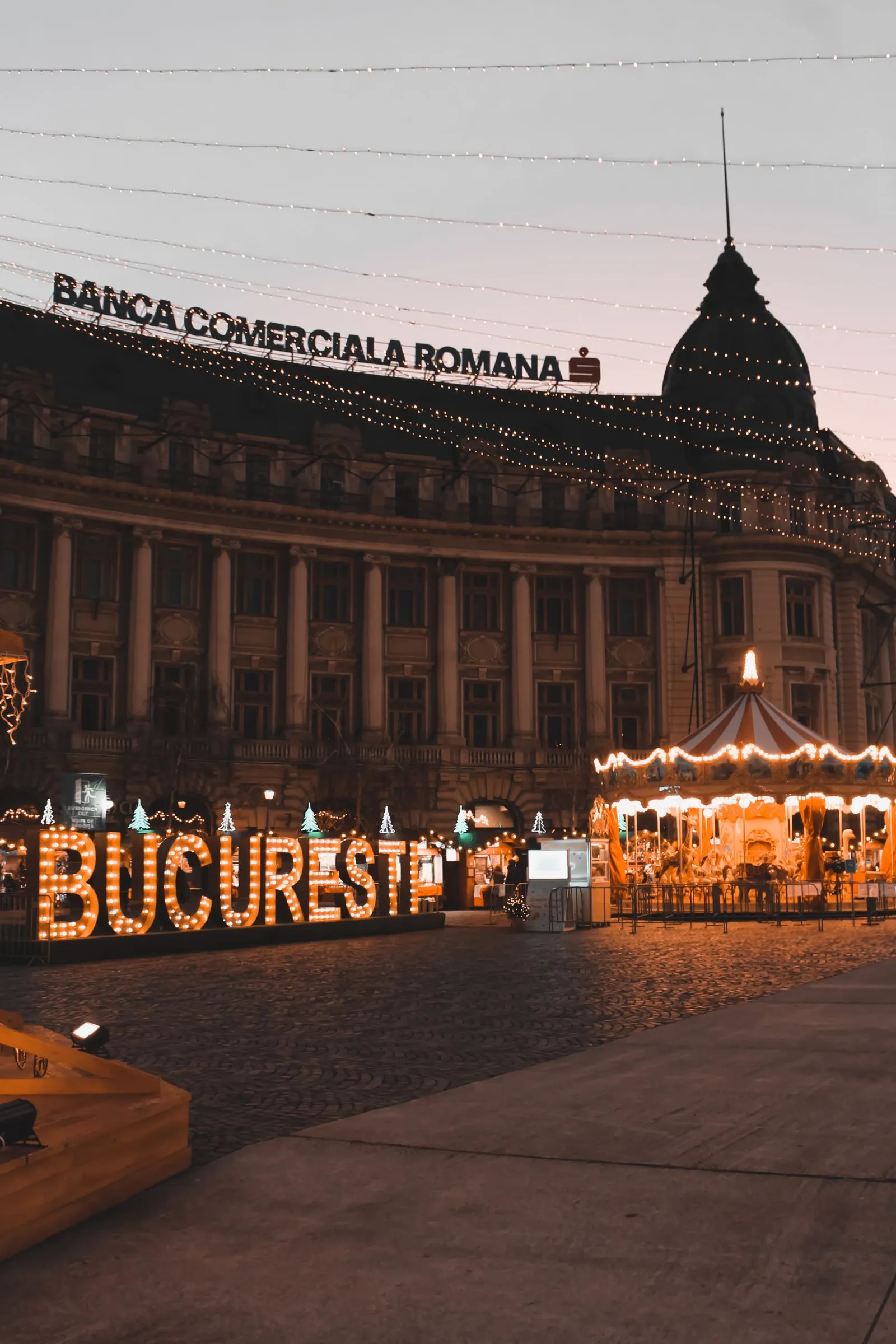Piața Universității din București seara, cu literele luminate BUCUREȘTI și un carusel de sărbători