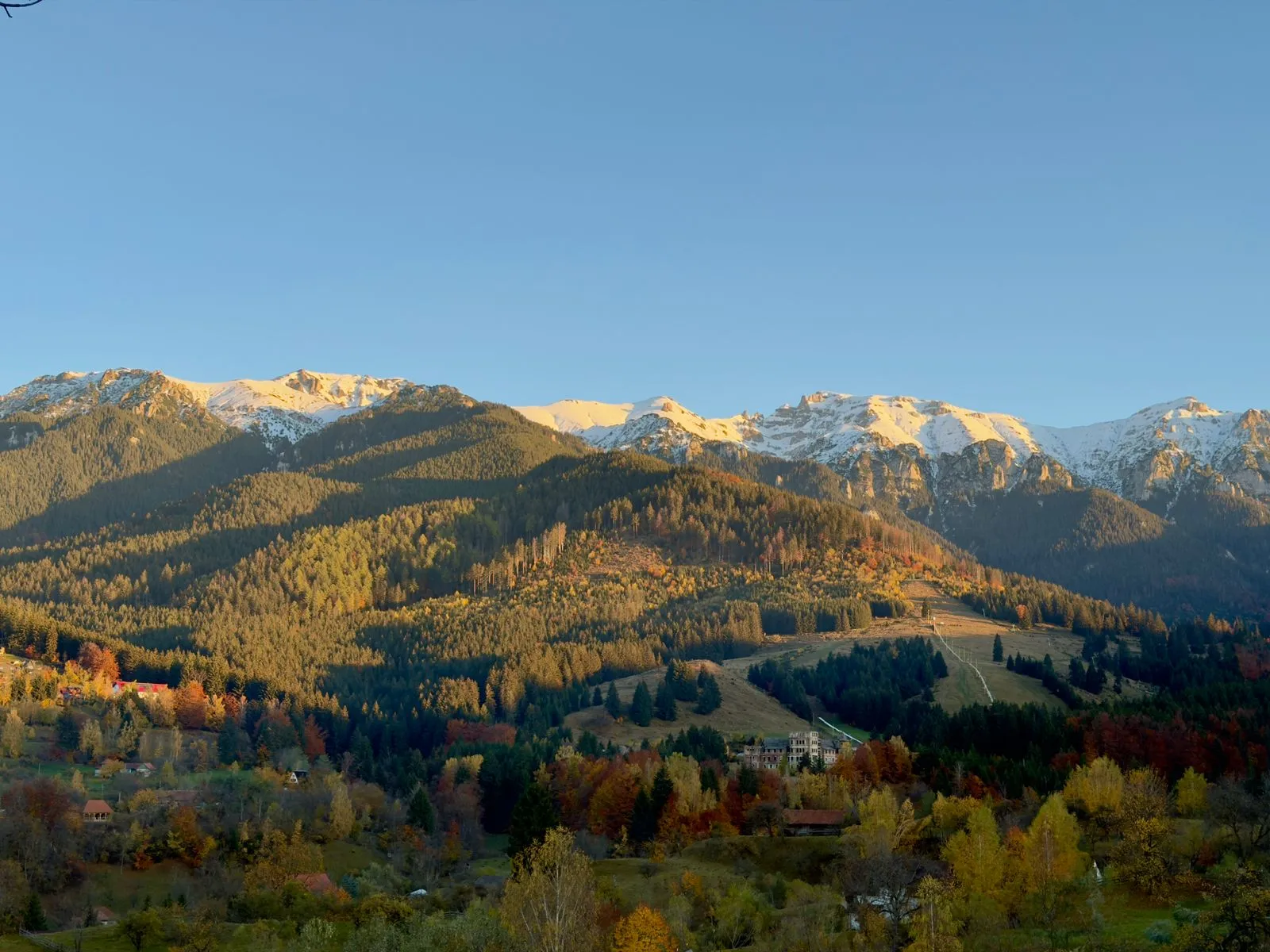Munții Bucegi toamna, cu păduri colorate deasupra Văii Prahovei și primele zăpezi pe creste