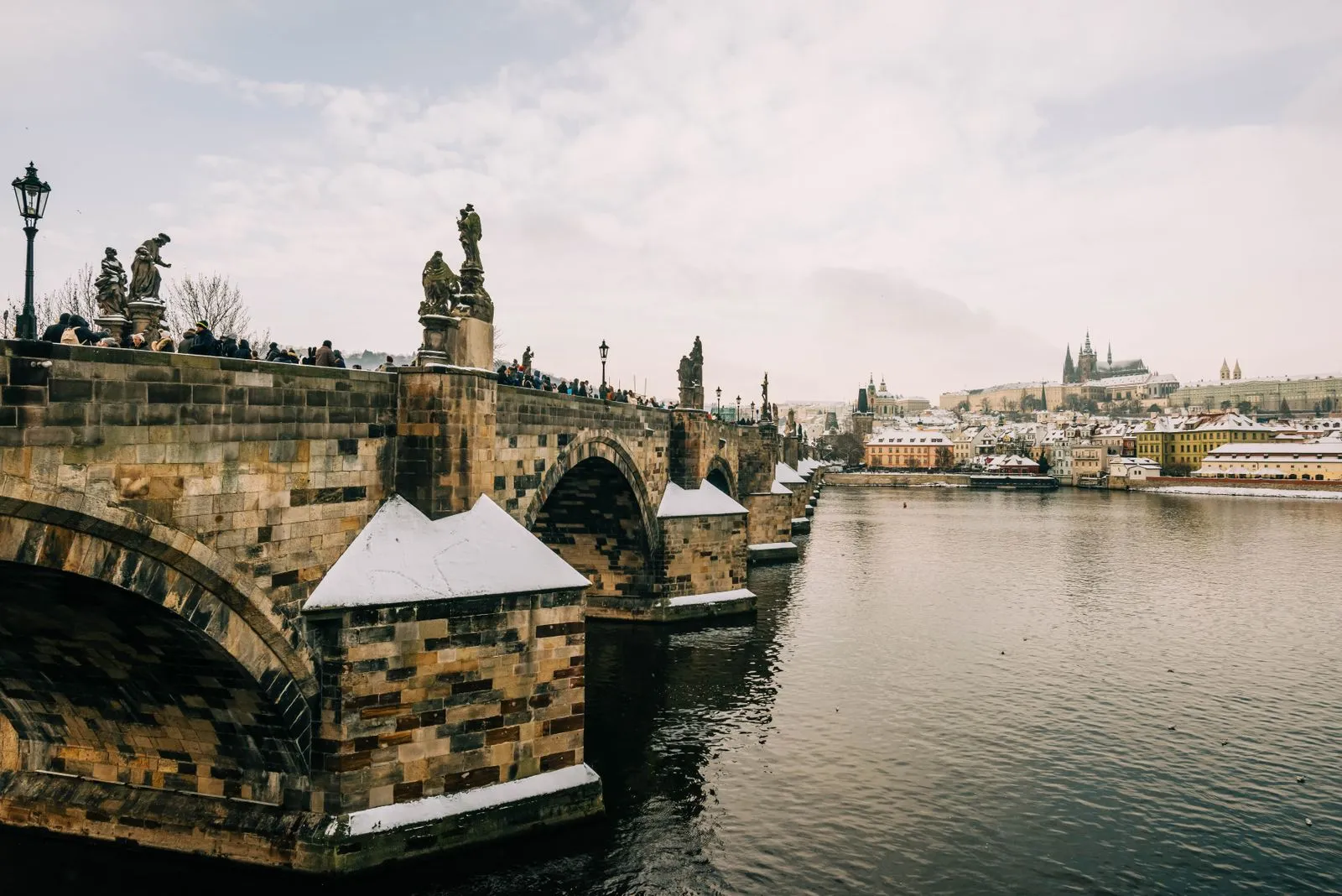 Podul Carol peste Vltava iarna, cu zăpadă pe parapete și Castelul Praga în depărtare