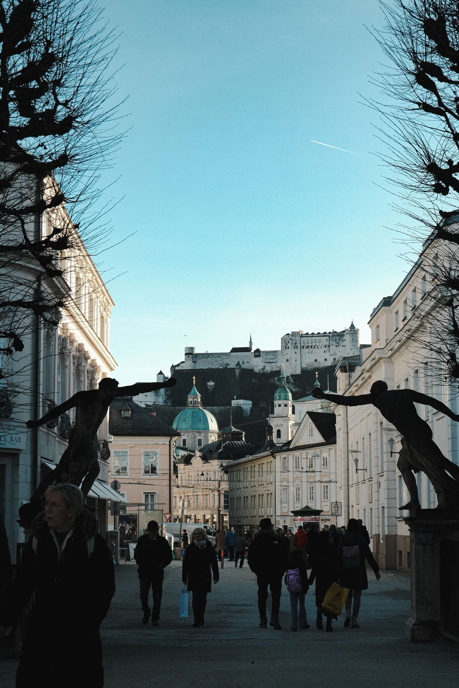 Oameni plimbându-se iarna pe o stradă din Salzburg, cu cetatea Hohensalzburg și cupolele din centrul vechi în fundal