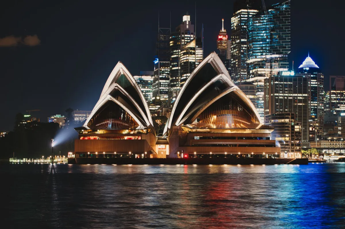 Opera din Sydney iluminată noaptea, cu zgârie-norii din centru reflectați în port